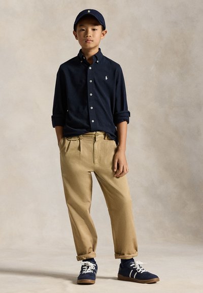 Bernliukas dėvi tamsiai mėlyną kepuraitę, tamsiai mėlyną marškinių su sagomis ir logotipu, smėlio spalvos kelnes bei tamsiai mėlynus sportbačius, stovintis su rankomis kišenėse prieš paprastą foną.