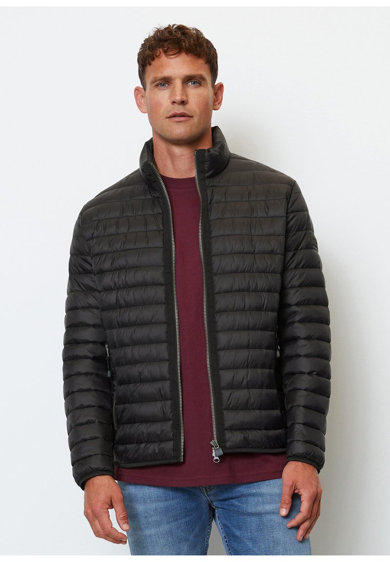 Chaqueta de plumas negra con un alto cuello, diseño acolchado, zipper frontal y bolsillos laterales, llevada sobre una camisa burdeos, exhibida sobre un fondo blanco.