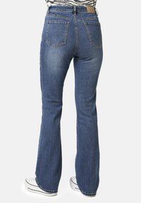 Persona con jeans de mezclilla acampanados azules y zapatillas blancas, vista desde atrás contra un fondo sencillo.