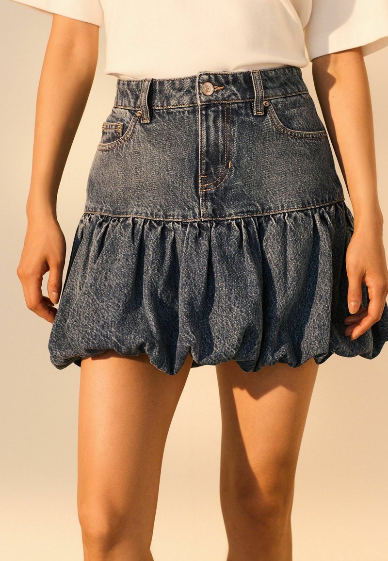 Fustă din denim cu talie ajustată și tiv volant, de culoare albastru deschis, cu detalii evidente de cusătură.