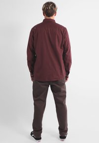 Chemise longue marron à manches longues avec poignet à bouton et un empiècement au dos. Associée à un pantalon marron et des baskets noires. Conception simple et décontractée.