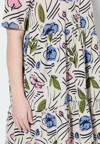 Kukallinen mekko beigen sävyissä, joissa on sinistä, vaaleanpunaista ja vihreää. Ominaisuuksiin kuuluu lyhyet hihat, rento malli ja kerroksellinen röyhelösuunnittelu.