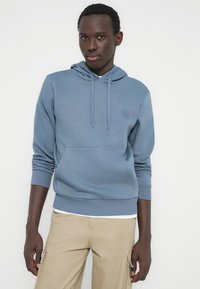 Ung mand iført en blå hættetrøje og beige cargobukser, der står foran en ensartet hvid baggrund.