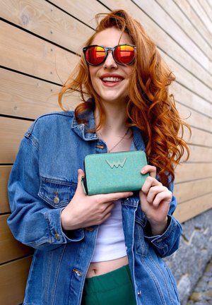 Teal textured wallet with a logo, held by a person in a denim jacket. Features red mirrored sunglasses and a wooden backdrop.