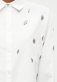 Λευκό πουκάμισο με κουμπιά, διακοσμημένο με ασημένιες ορθογώνιες χάντρες διάσπαρτες στο μπροστινό μέρος και στο μανίκι, με κλασικό γιακά.