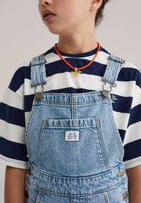 Salopetă din denim cu bretele reglabile, de culoare albastru deschis, cu buzunare frontale. Poartă peste un tricou cu dungi albastre și albe, accesorizat cu un colier roșu cu mărgele.