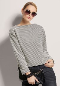 Gestreiftes Langarmoberteil in Creme und Schwarz, kombiniert mit einer glänzenden schwarzen Handtasche. Die Accessoires umfassen ein geschichtetes schwarzes Armband und eine Sonnenbrille.