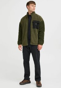 Olive green fleece jacket with a black chest pocket and zip closure, paired with dark pants and brown patterned footwear.