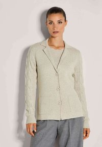 Frau in beiger, geknöpfter Strickjacke mit Kabelstrick-Ärmeln und zwei Fronttaschen, kombiniert mit lockeren grauen Hosen.