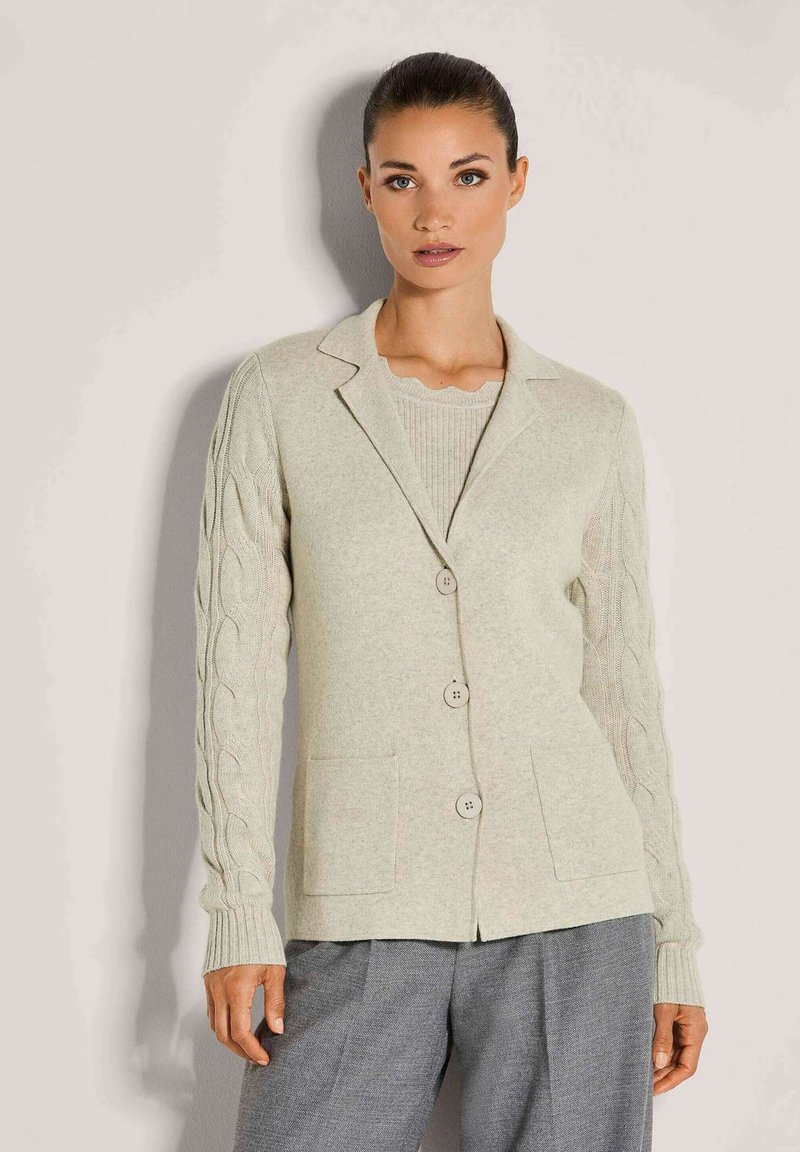 Frau in beiger, geknöpfter Strickjacke mit Kabelstrick-Ärmeln und zwei Fronttaschen, kombiniert mit lockeren grauen Hosen.