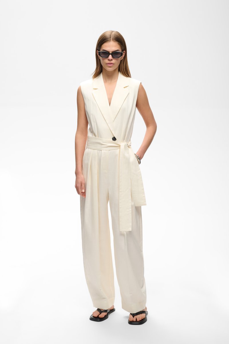 Femme portant une combinaison crème sans manches avec ceinture, lunettes de soleil noires et sandales noires à bout ouvert, debout devant un fond blanc uni.