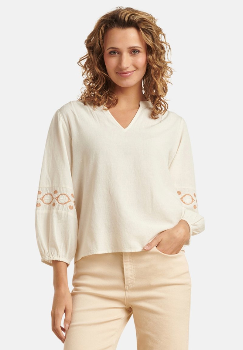 Cremefarbige Bluse mit V-Ausschnitt und Ballonärmeln, die am Saum mit rustfarbenen Stickereien verziert ist. Weicher, strukturiert Stoff.