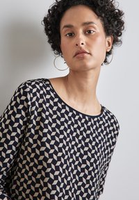 Navy toppi, jossa on geometrinen kuvio vaalean kerman ja vaaleanharmaan sävyissä, pyöreällä pääntiellä ja pitkillä hihoilla. Sileä kangas, jossa on minimalistinen muotoilu.