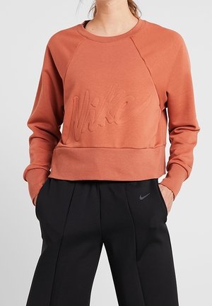 Frau trägt ein rostfarbenes Nike-Sweatshirt mit geprägtem Logo und schwarze Nike-Hose, die Hände in den Taschen, vor einfarbigem Hintergrund.