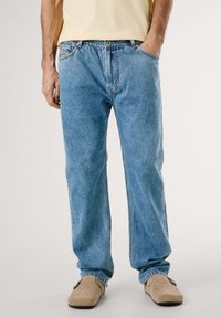 Jeans de mezclilla de lavado claro con un corte relajado, diseño de cinco bolsillos y costuras visibles. Llevados con zapatos beiges de fácil puesta.
