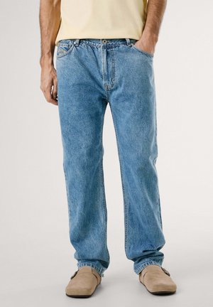Jeans en denim bleu clair avec une coupe décontractée, un design à cinq poches et des coutures visibles. Portés avec des chaussures à enfiler beiges.