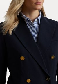 Marineblauwe dubbelgevoerde blazer met gouden knopen, met een scherp design van de revers. Daaronder draagt de persoon een blauw-wit gestreept shirt.