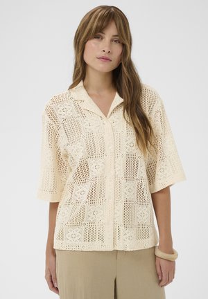 Vrouw draagt een crèmekleurig gehaakt kortärmelig overhemd met knopen en beige broek, staat met een neutrale gezichtsuitdrukking tegen een witte achtergrond.
