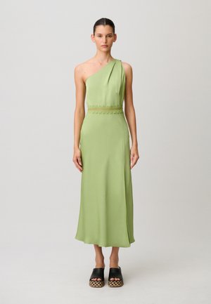 Mujer joven de pie, con un vestido midi verde claro de un solo hombro y cintura ajustada, y sandalias negras de plataforma.