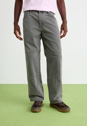 Mann trägt verblasste graue gerade geschnittene Jeans und braune Wanderschuhe, steht auf grünem Boden vor weißem Hintergrund.