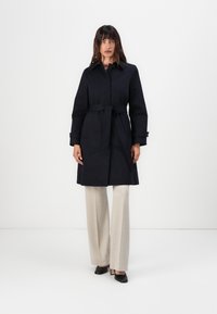 Marinefarbener trenchcoat mit Gürtel, Kragen, langen Ärmeln, Seitentaschen und gerader Schnitt. Getragen über beigen weiten Hosen und schwarzen Absatzschuhen.