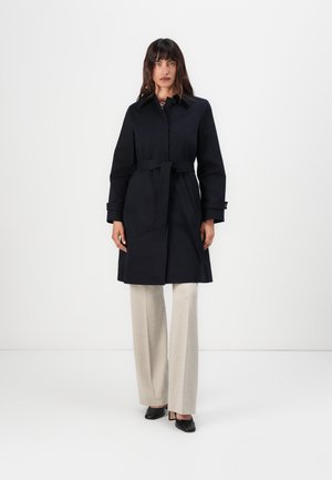 Manteau trench ceinturé bleu marine avec un col, des manches longues, des poches latérales et une coupe droite. Porté sur un pantalon large beige et des talons noirs.