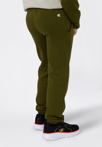 Person i olivgröna joggingbyxor med Champion-logotyp nära fickan och svarta träningsskor med vita sulor och orange detaljer.