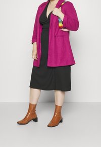 Fuchsia blazer over een zwarte jurk met een diepe halslijn. Bruine enkellaarsjes met een vierkante neus en lage hakken. Kleurrijke geweven schouderband.