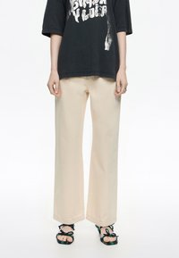 Camiseta negra de gran tamaño con gráfico blanco, pantalones anchos beige y sandalias negras con acentos tejida coloridos. Texturas de tela suaves visibles.