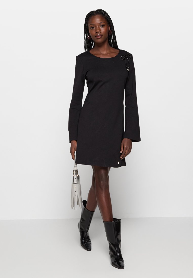 Czarna sukienka z długimi rękawami i rozszerzanymi mankietami, z akcentem w postaci kokardy z przodu, w połączeniu z błyszczącymi czarnymi kozakami do kolan i jasną torebką.