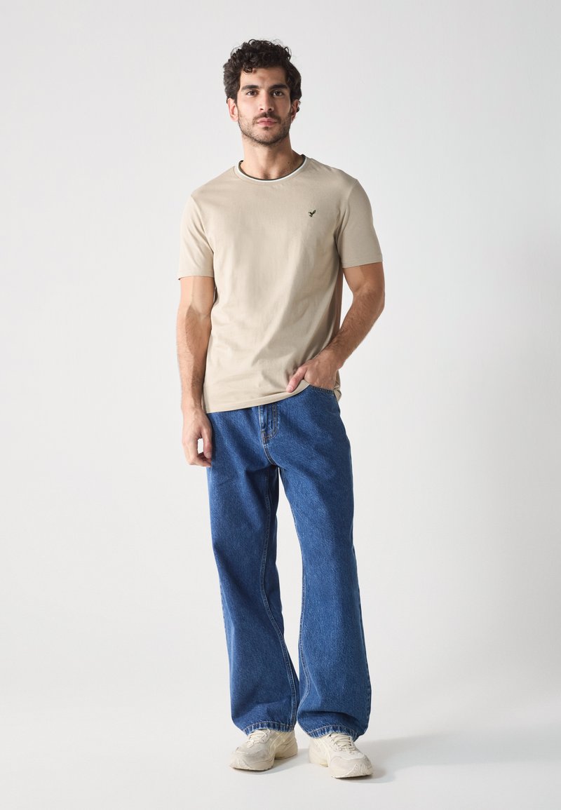 Camiseta de algodón color beige con cuello redondo y logo verde, combinada con jeans de mezclilla azul de corte holgado, acompañada de zapatillas de color claro.
