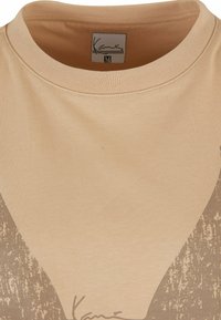 Beige bomulls skjorte med rund hals, med et brunt grafisk mønster og en trykt signatur nederst. Størrelseslabel: M.