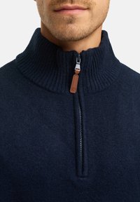 Marineblå strikket sweater med høj ribkant, halv lynlås lukning og en brun læderdetalje på lynlås-trækkeren. Blød tekstur.