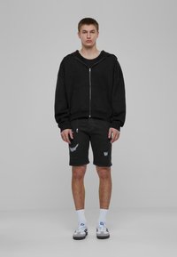 Juodas zip-up džemperis su kengūrų kišene, derinamas su nusidėvėjusiomis juodomis šortais. Modelis dėvi baltas kojines ir pilkas sportines batus.