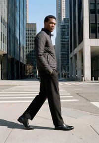 Jeune homme portant une veste noire matelassée et un pantalon noir marchant sur le trottoir en ville près d'un passage piéton entre de hauts bâtiments.