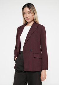 Bordeaux blazer med skræddersyet design, enkelt knaplukning, klapslag og én lomme. Båret over en hvid skjorte med mørke bukser.