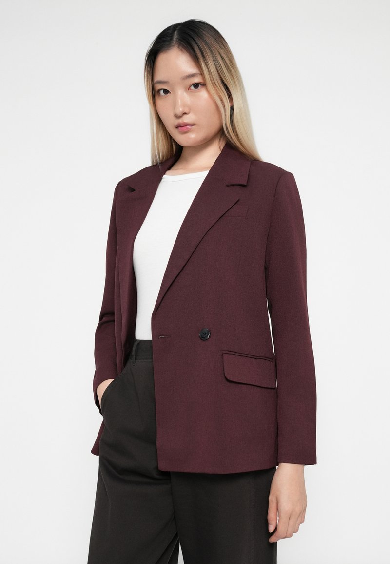 Blazer bordeaux con un design sartorial, chiusura a un bottone, colletto a scollo e una tasca. Indossato sopra una camicia bianca con pantaloni scuri.