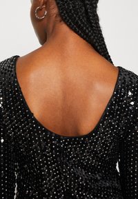 Czarna sukienka z cekinami, z niskim dekoltem na plecach, długimi rękawami oraz fakturowanym materiałem o błyszczącej powierzchni.