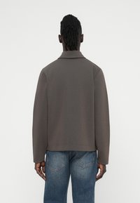 Καφέ, υφασμάτινο μπουφάν με γιακά και άνετη εφαρμογή, συνδυασμένο με τζιν ανοιχτού γαλάζιου χρώματος. Η πλάτη έχει λείο ύφασμα με διακριτικές ραφές.