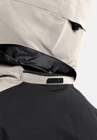 Giacca beige e nera con tasca con zip, chiusura in velcro e un tessuto testurizzato e resistente all'acqua. Presenta un design elegante e moderno.