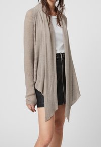 Cárdigan beige de canalé con apertura frontal, mangas largas y diseño drapeado. Lleva puesto sobre una camiseta blanca, combinado con una falda negra.