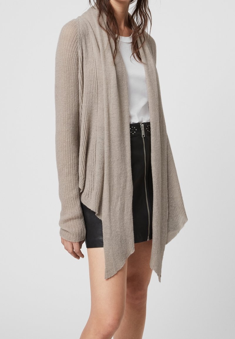 Cárdigan beige de canalé con apertura frontal, mangas largas y diseño drapeado. Lleva puesto sobre una camiseta blanca, combinado con una falda negra.