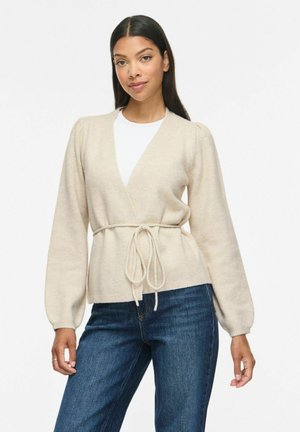 Femme aux longs cheveux foncés portant un pull beige cache-cœur noué à la taille sur un top blanc et un jean bleu, debout devant un fond blanc.