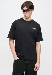 Μαύρο κοντομάνικο T-shirt από λείο ύφασμα, με μικρό λευκό λογότυπο στο αριστερό στήθος και χαλαρή εφαρμογή. Φορεμένο με σκούρο τζιν παντελόνι.