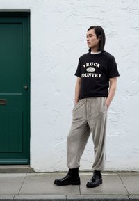 Juoda medvilninė marškinėliai su užrašu „TRUCK COUNTRY“, smėlio spalvos korduro kelnės, juodi batai, stovintys priešais tekstūruotą baltą sieną ir žalią duris.