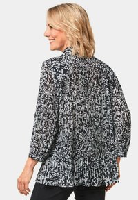 Zwart-wit gemusterde blouse met geplooide textuur, lange mouwen, hoge kraag en een losse pasvorm. Bevat abstracte ontwerpen in verschillende vormen.