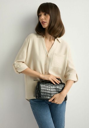 Kobieta o brązowych włosach do ramion, ubrana w beżową koszulę na guziki i niebieskie dżinsy, trzymająca czarną, teksturowaną torebkę na tle białej ściany.