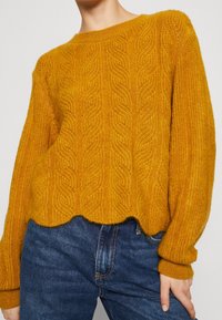 Senfgelbes Kabelstrickpullover mit gewelltem Saum, getragen mit dunkelblauen Denim-Jeans von einer stehenden Person.