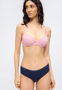 Γυναίκα που φοράει ροζ σουτιέν με δαντέλα και μπλε σκούρο εσώρουχο με δαντέλα, κοιτάζοντας προς τα δεξιά της σε απλό φόντο.