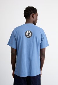 Tricou din bumbac albastru, cu mâneci scurte, având un logo alb-negru într-un design circular pe spate. Conic clasic, cu o textură moale.